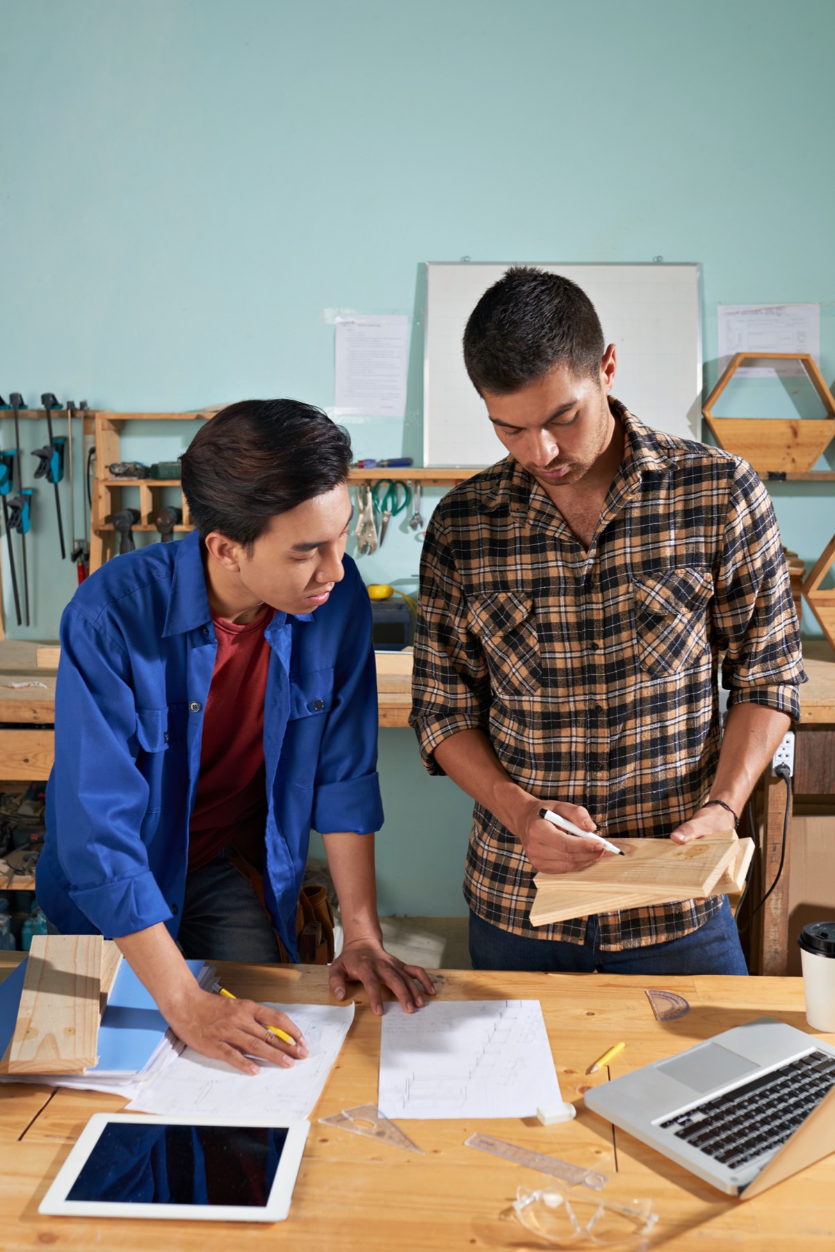 Two people at a workbench checking a wooden part against a drawing.